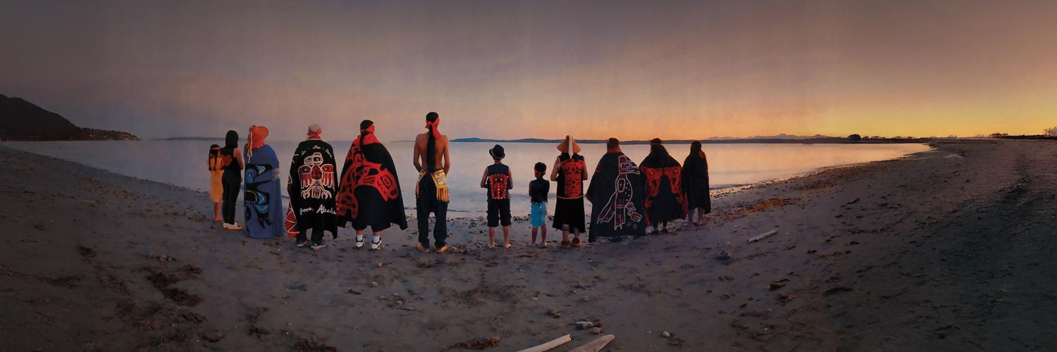 Native Americans dressed in traditional clothing standing on the beach watching the sunrise.