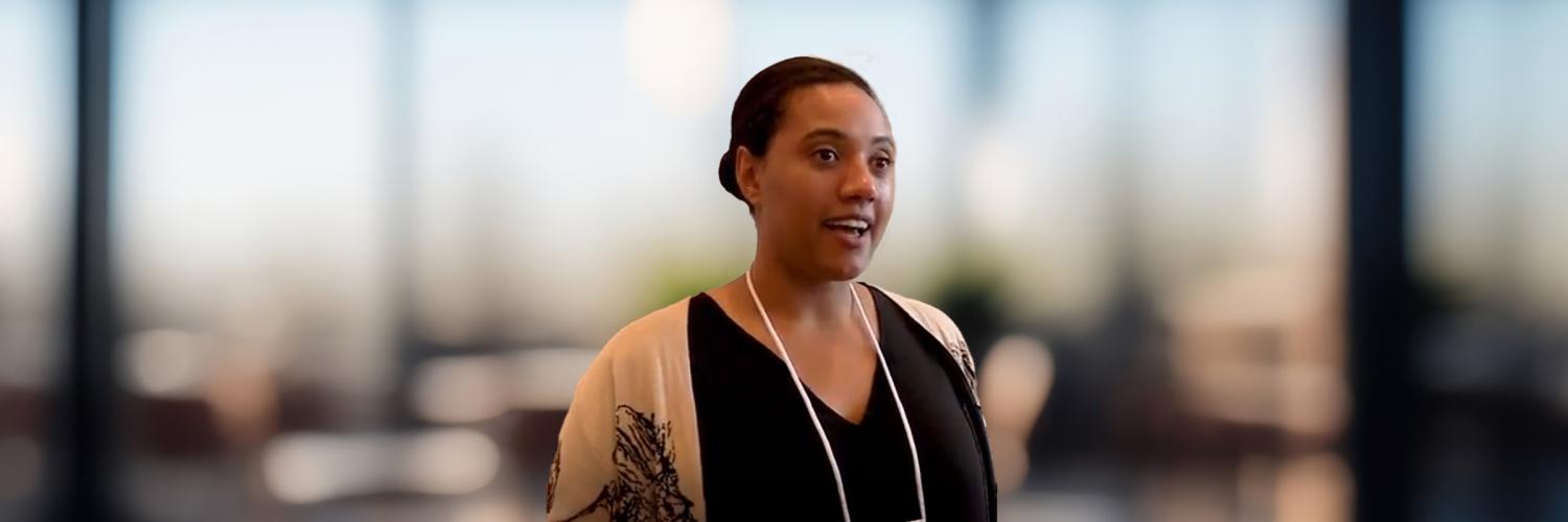 Photo of a community worker, Leah, standing indoors with a blurry background of tall window panes.