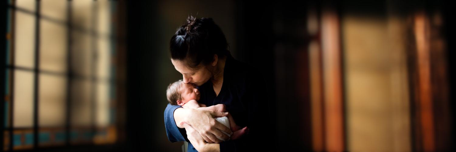 A photo of a mother holding her newborn close in both hands. They are indoors with a few closed windows behind them with daylight glowing behind.