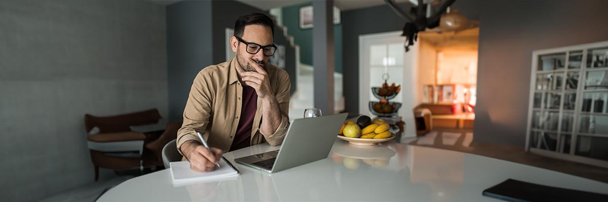 A man looking at a laptop and writing in a notebook