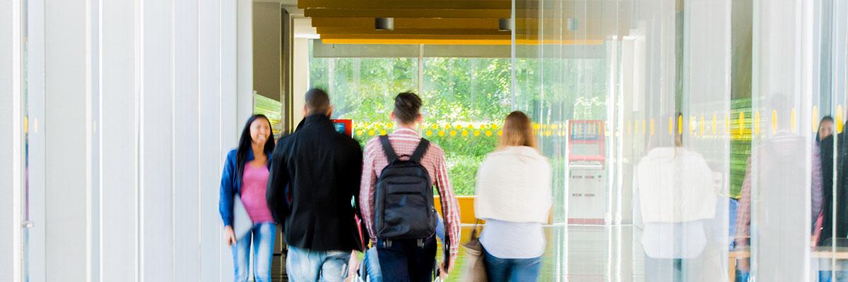 College student walking down a bright hallway with floor to ceiling windows on either side.