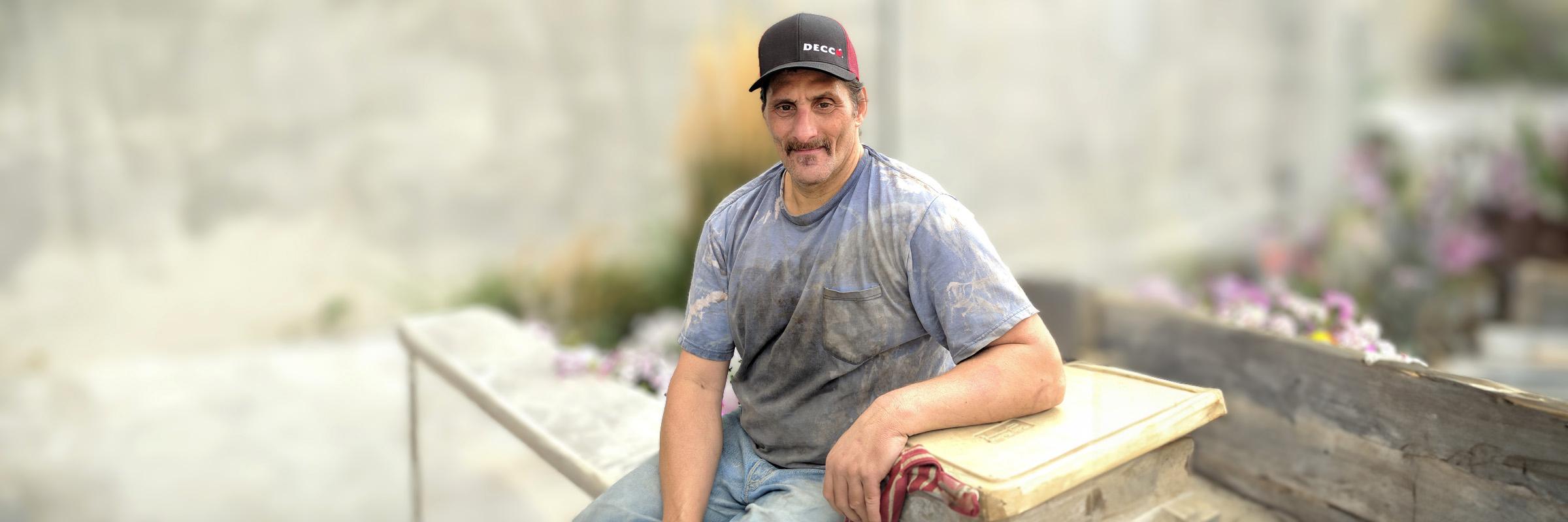 A male is smiling and sitting on a wooden bench with his arm leaning on a box. There is a blurred concrete and wooden flowerbed behind him. 