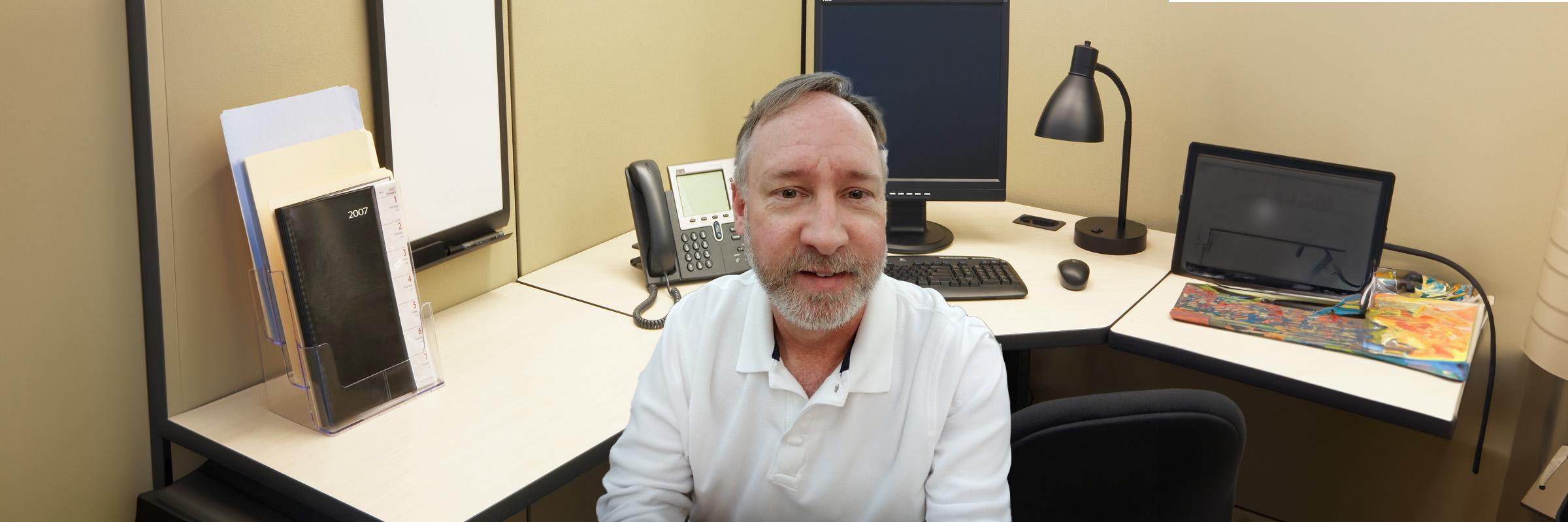 A photo of a male in a white shirt sitting in an office cubicle.