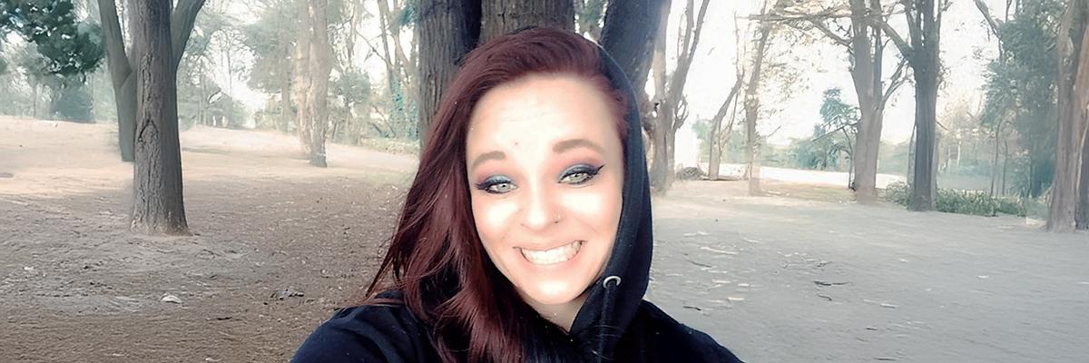A selfie photo of a woman smiling in the middle of forest with many trees sprawled around. 