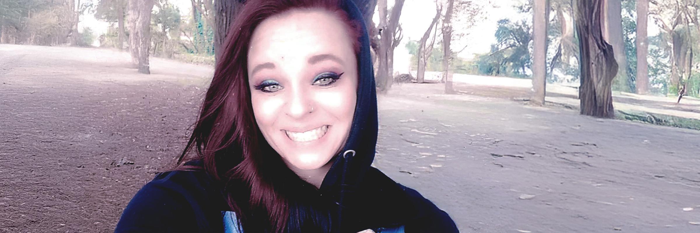 A selfie photo of a woman smiling in the middle of forest with many trees sprawled around. 