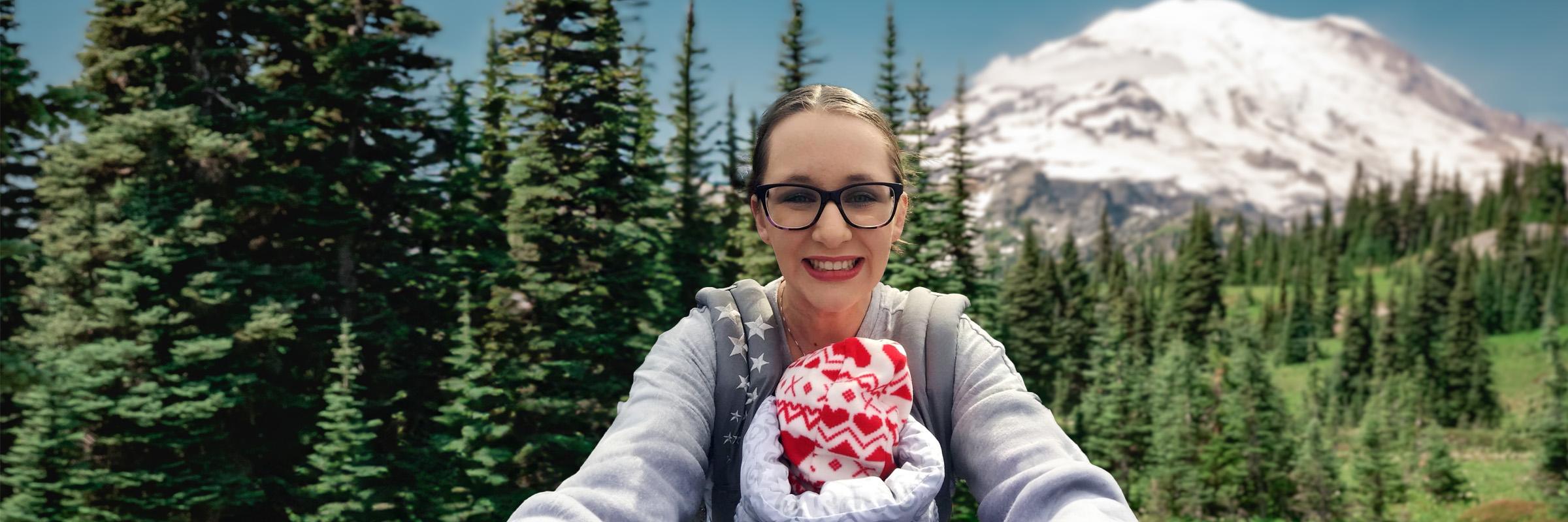 Photo of a mother carrying her infant daughter with a front facing baby carrier. Behind them is a tree line and a snowy mountain with blue skies. 