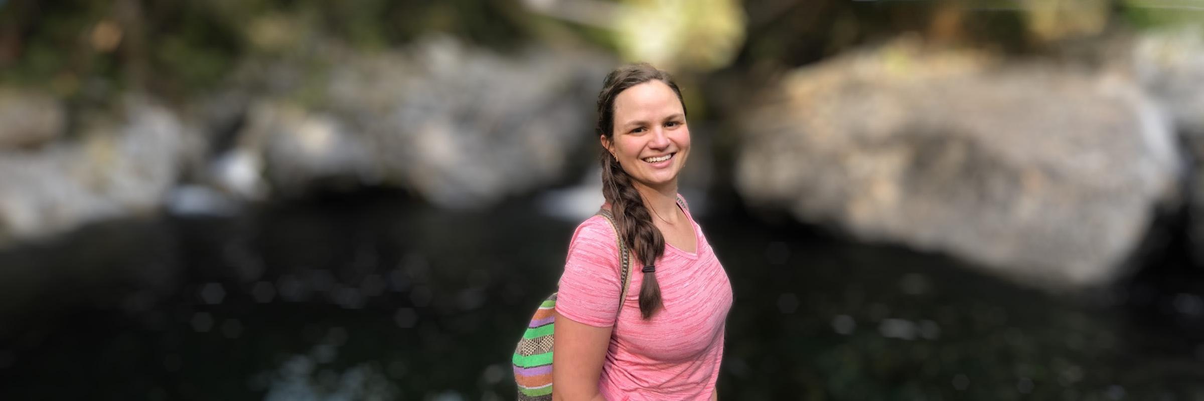 Photo of a young adult female standing outside hiking in front of two boulders. 