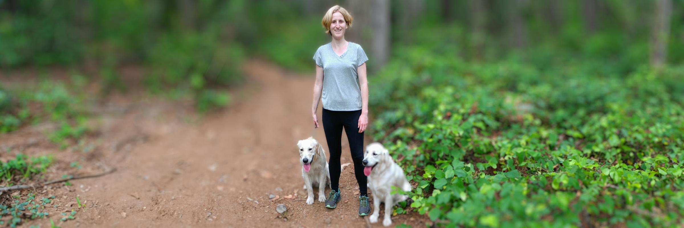 Photo of a smiling female person, happily standing in a green forest walking trail with 2 white dogs sitting beside her. 