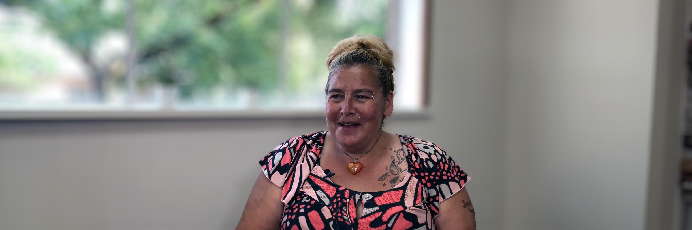 Photo of a female client smiling and sitting down indoors with a window behind her.