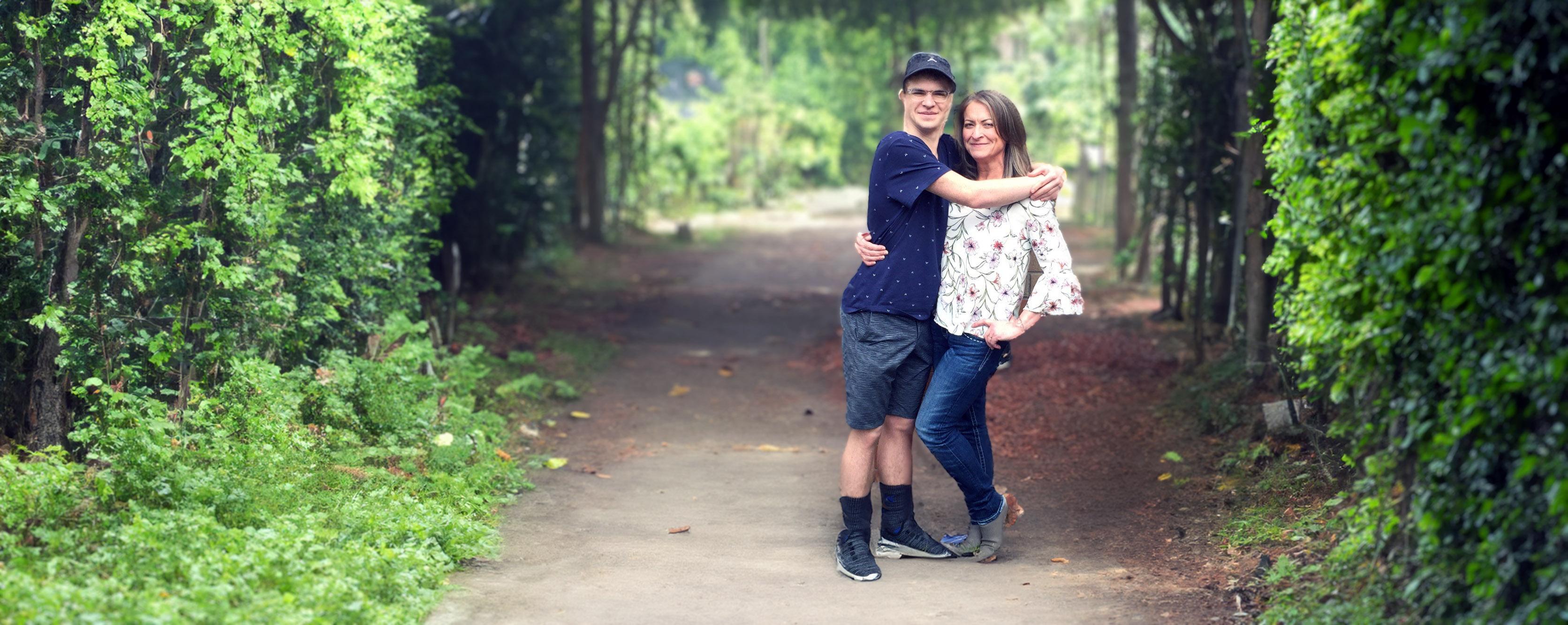 Photo of a mother and son hugging each other along a tree covered path.