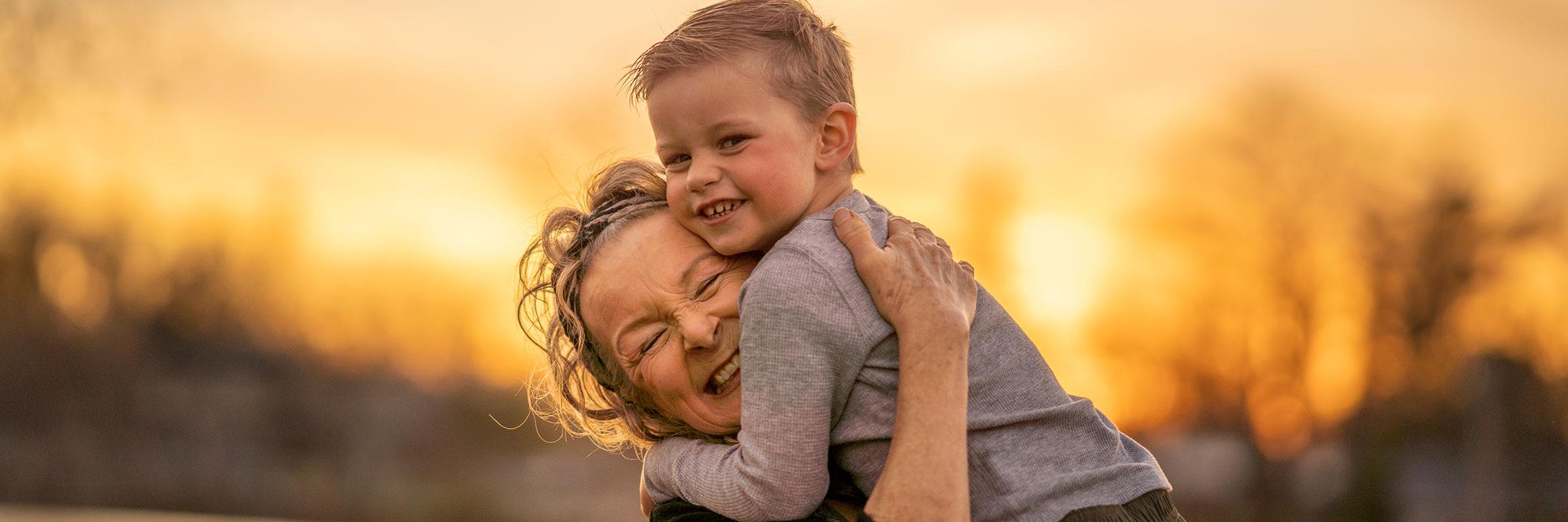 A photo of a grandmother and her grandson smiling outside in the forest under a sunset blurred background.