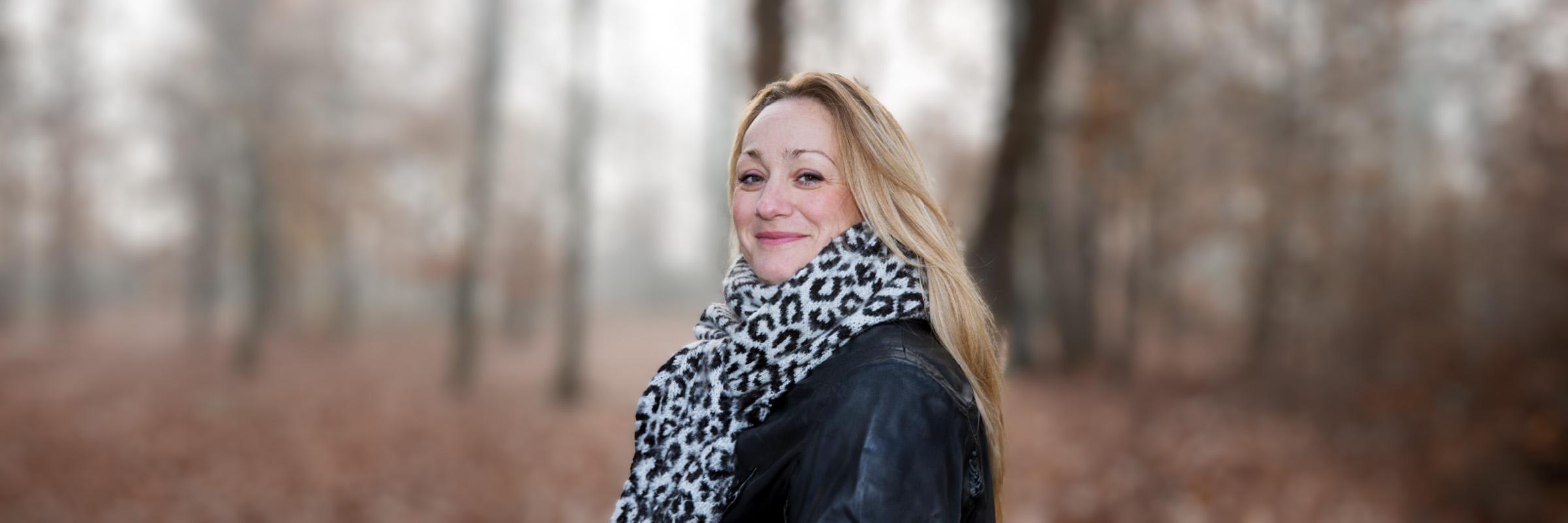 Photo of a woman with a fur scarf and black leather jacket smiling. She is standing standing in the woods during a cloudy day.