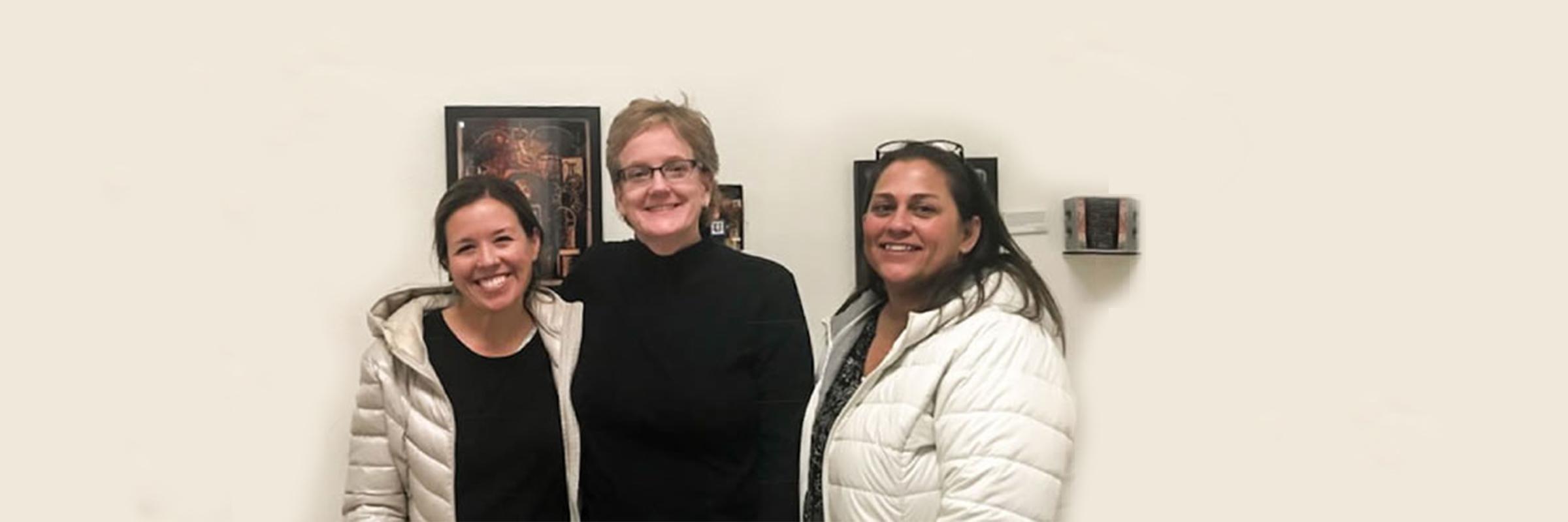 Amy Blondin, Deb Morris, and Karin Kramer standing in front of artwork, looking at the camera and smiling