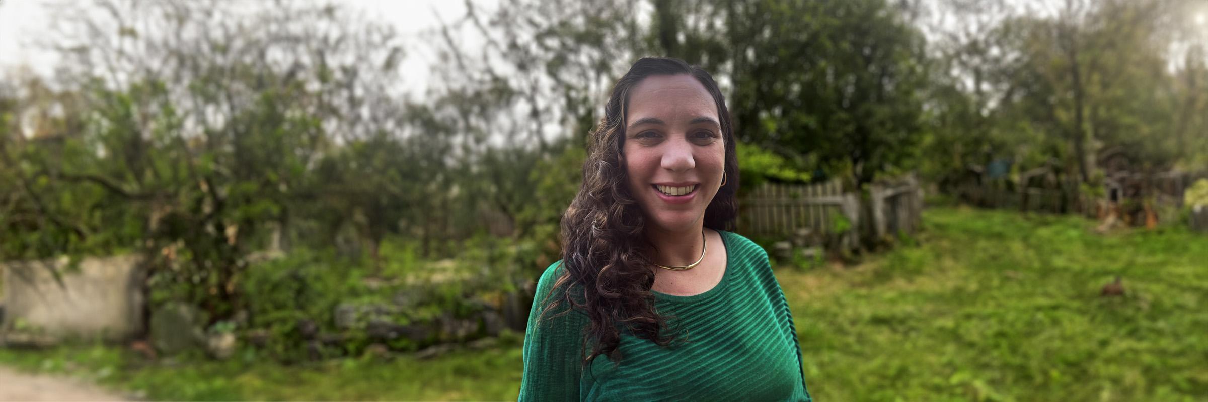 Photo of a female wearing a green and black striped sweater standing outdoors in a park with green foliage in the background. 